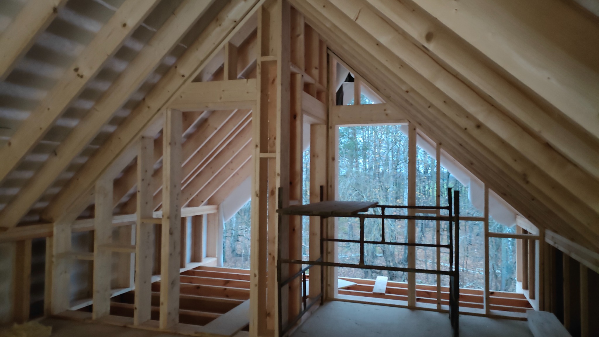 Wnętrze w trakcie budowy z drewnianą konstrukcją szkieletową poddasza, widoczne rusztowanie i okna z widokiem na las.