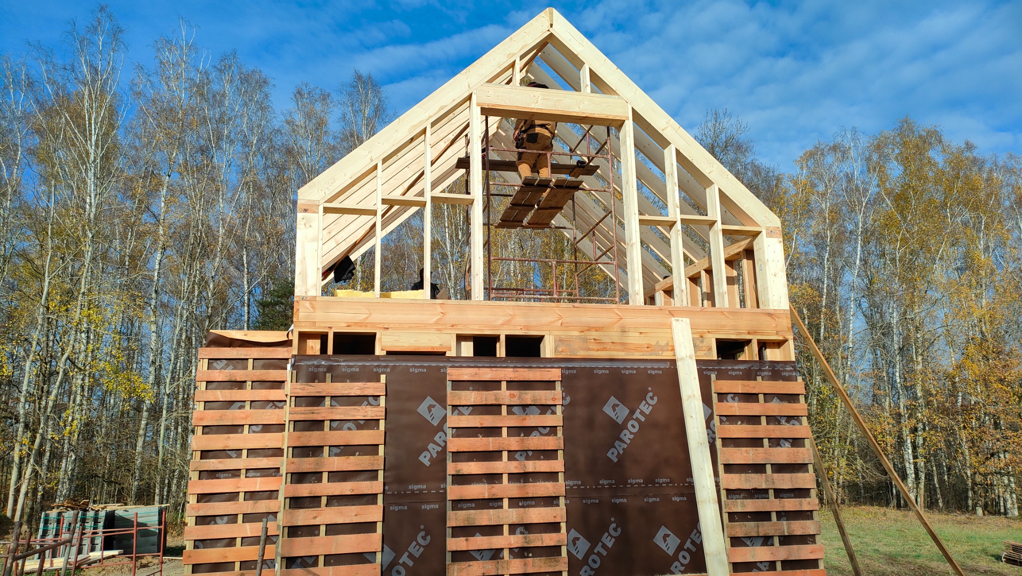 Wznoszenie drewnianej konstrukcji domu szkieletowego na fundamencie obłożonym brązową folią izolacyjną, widoczny pracownik na rusztowaniu i las brzozowy w tle.