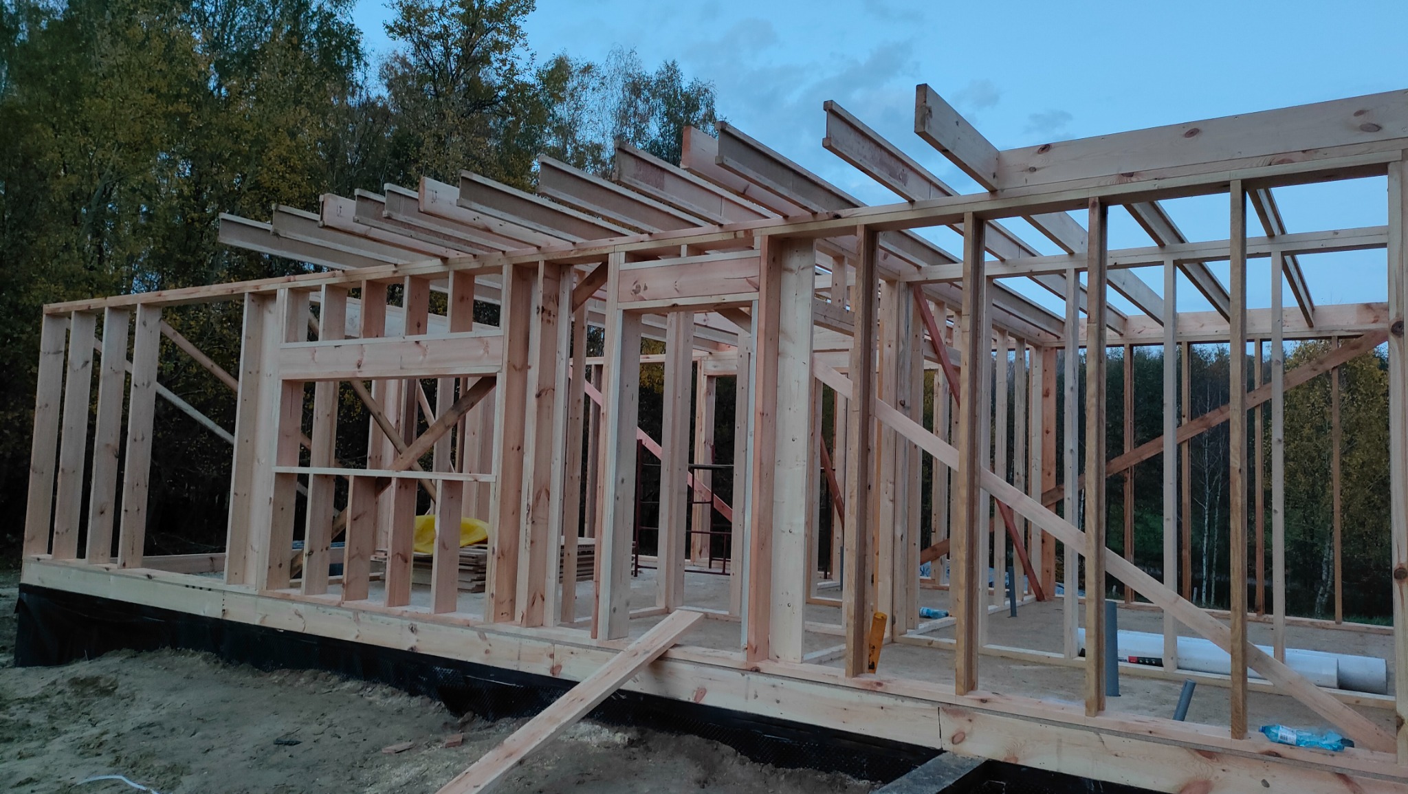 Wznoszenie konstrukcji domu z drewnianych belek na fundamencie, widoczne elementy szkieletu ścian i dachu, w tle las.