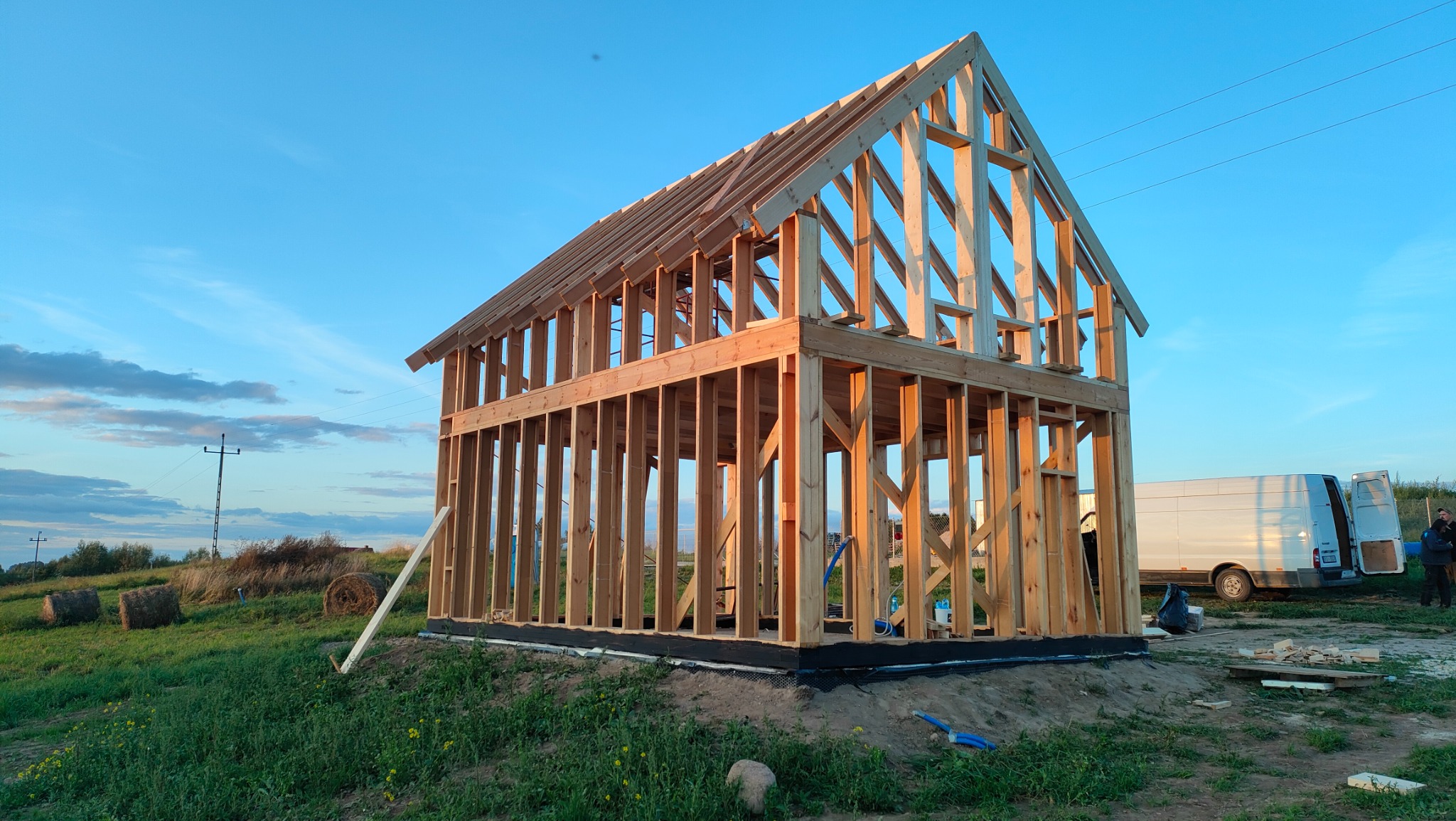Wznoszenie konstrukcji szkieletowej domu z drewnianych belek na tle błękitnego nieba, widoczny biały van i bele siana na łące.