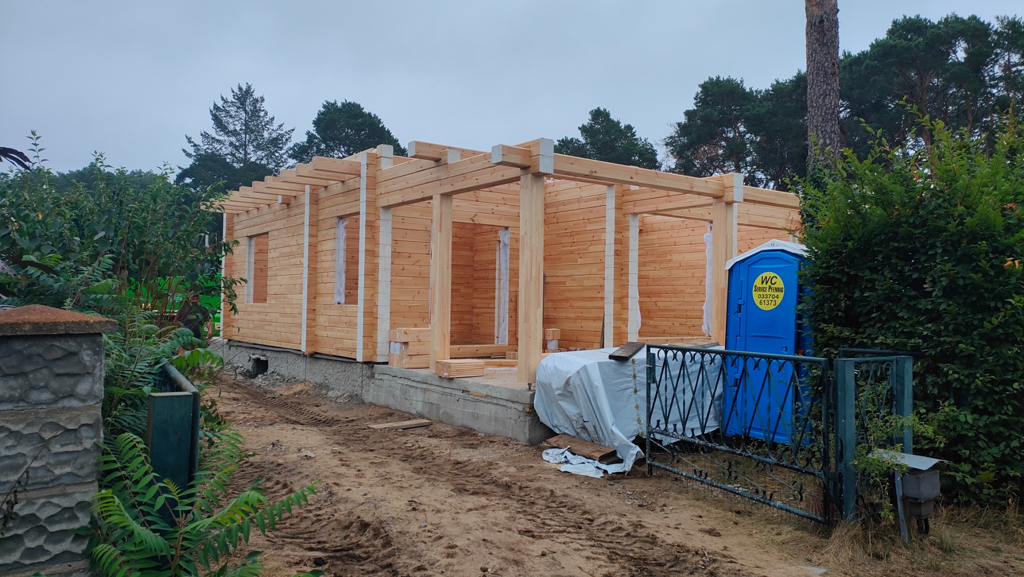 Surowa konstrukcja drewniana budynku z widoczną ramą ścian i dachu, na zewnątrz przenośna toaleta.