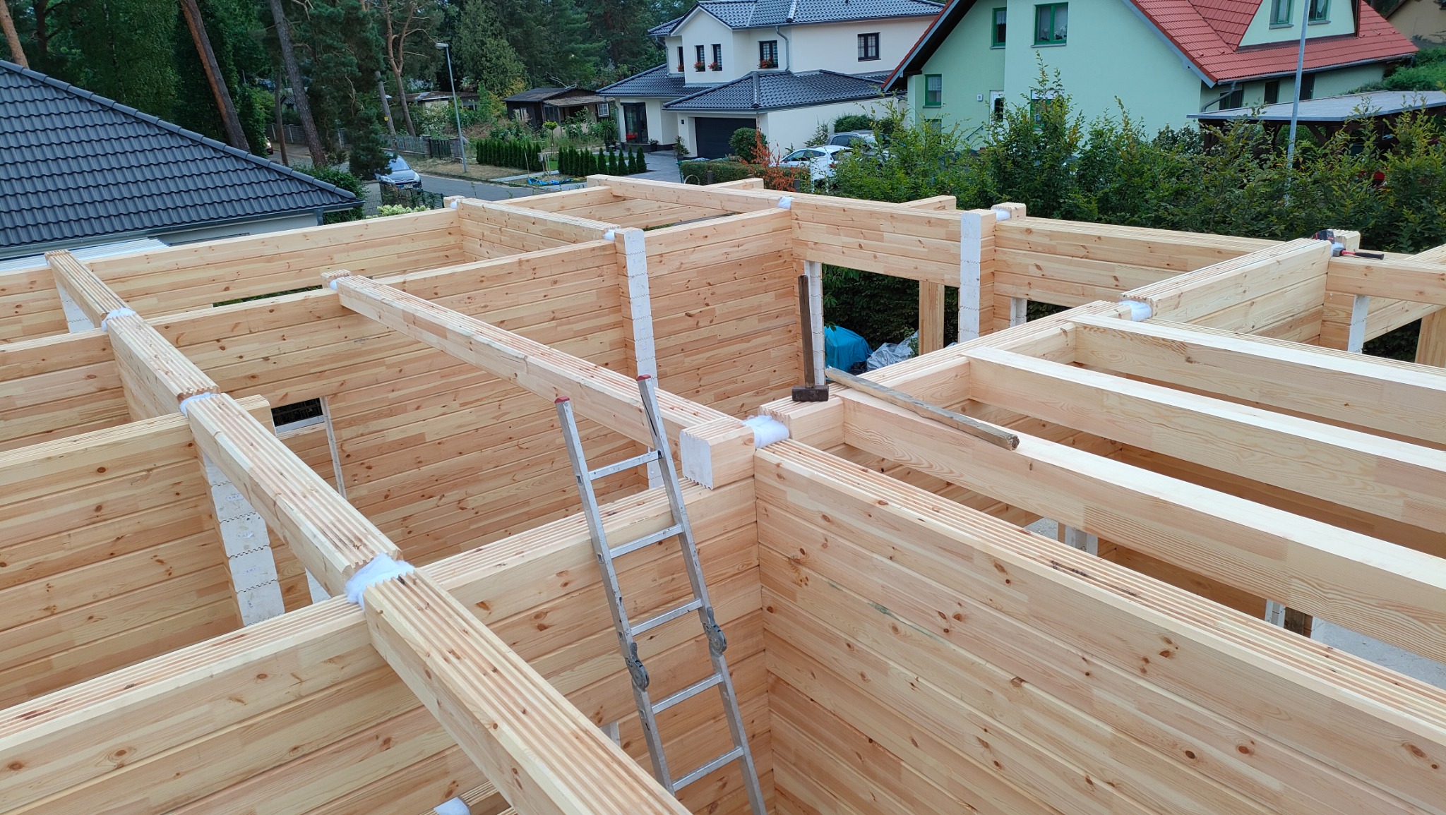 Konstrukcja ścian i stropu z drewna klejonego warstwowo podczas budowy domu, drabina oparta o ścianę, widoczne domy w tle.