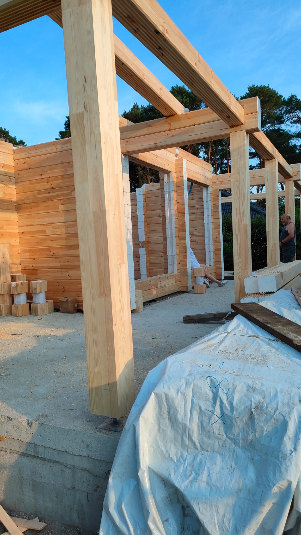 Wznoszenie konstrukcji budynku z drewnianych belek, widoczne jasne drewno, niebieskie niebo w tle, na pierwszym planie plandeka z niebieskimi oznaczeniami.