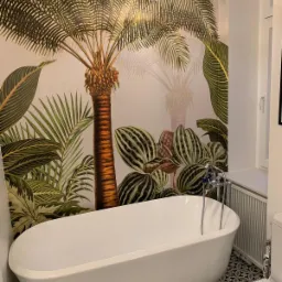 Biała wanna wolnostojąca na tle ściany z fototapetą przedstawiającą tropikalną roślinność, oświetlona wiszącą lampą z bursztynowym kloszem. W tle widoczna toaleta i grzejnik.