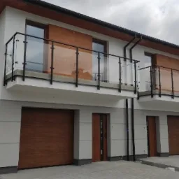 Dwa nowoczesne, bliźniacze domy z balkonami zabezpieczonymi czarnymi, szklanymi konstrukcjami, z drewnianymi akcentami na elewacji i bramach garażowych.