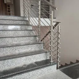 Schody wyłożone szarym kamieniem z nowoczesną metalową balustradą ze stali nierdzewnej z cienkimi prętami i drewnianą drabinką w tle.