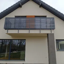 Szara balustrada balkonowa z poziomych profili na tle jasnej elewacji domu z ciemnoszarą stolarką okienną i dachem.