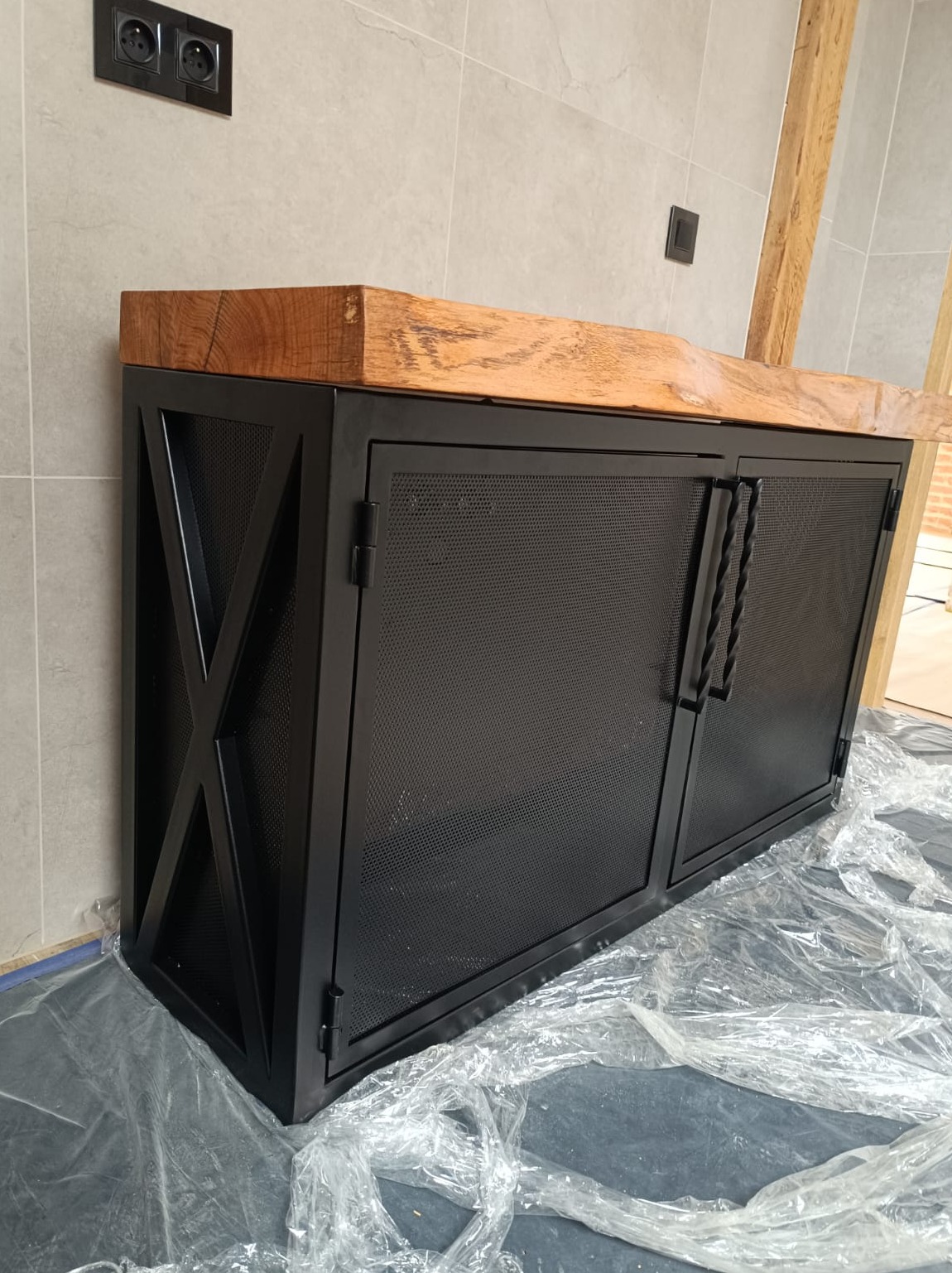 Metalowa szafka w stylu industrialnym z drewnianym blatem, czarne, perforowane drzwiczki, dekoracyjne uchwyty, widoczne zabezpieczenia podłogi.