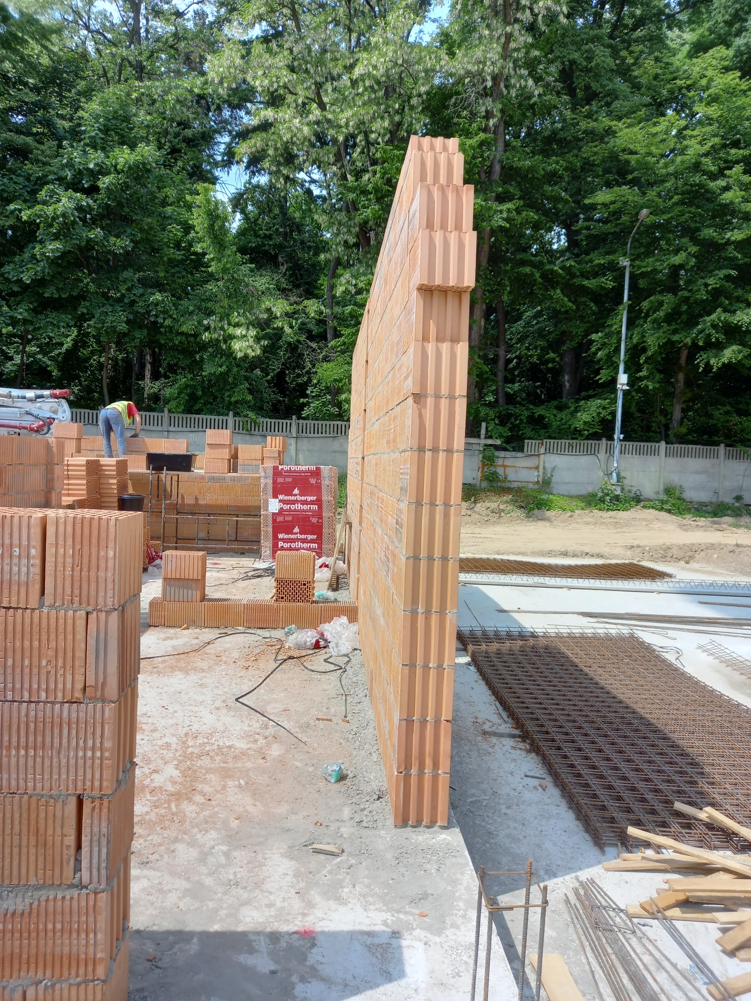 Wznoszenie ściany z pustaków ceramicznych Porotherm na budowie domu, widoczne zbrojenie fundamentów i pracownik w tle.