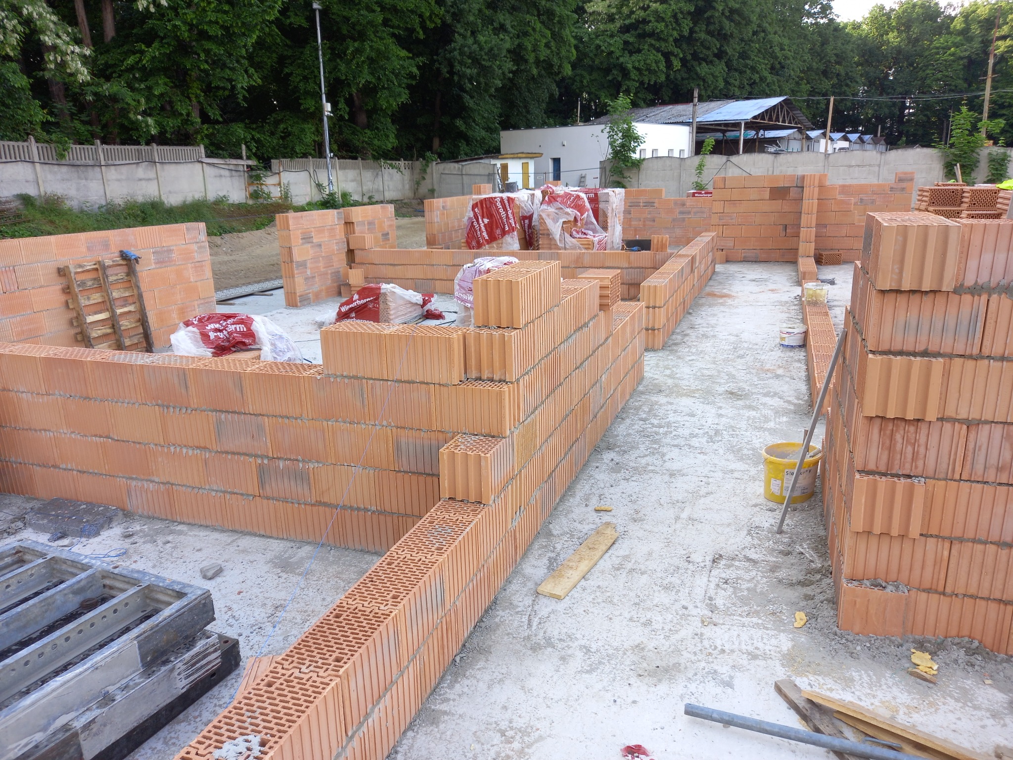 Fundamenty domu z cegły ceramicznej Porotherm w trakcie murowania, widoczne palety z cegłami, żółte wiadro i elementy szalunku.
