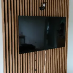 Drewniana ściana z pionowych listew, z zamontowanym telewizorem i lampą ścienną w stylu industrialnym, widoczne gniazdka elektryczne i przewody.