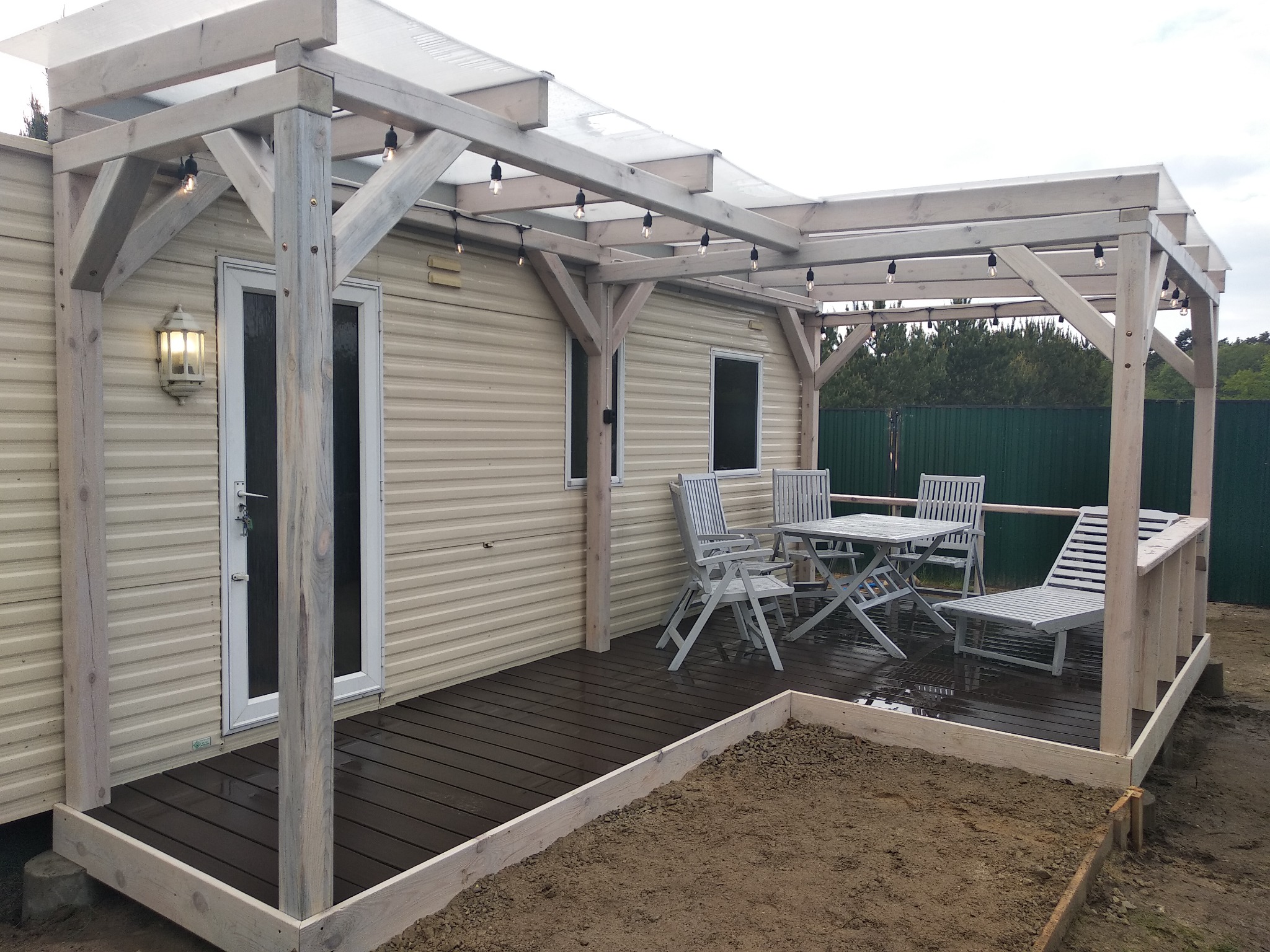 Zadaszony taras z drewnianą konstrukcją, oświetleniem żarówkowym i meblami ogrodowymi, przylegający do domu z sidingiem, z przygotowanym miejscem na ogródek.