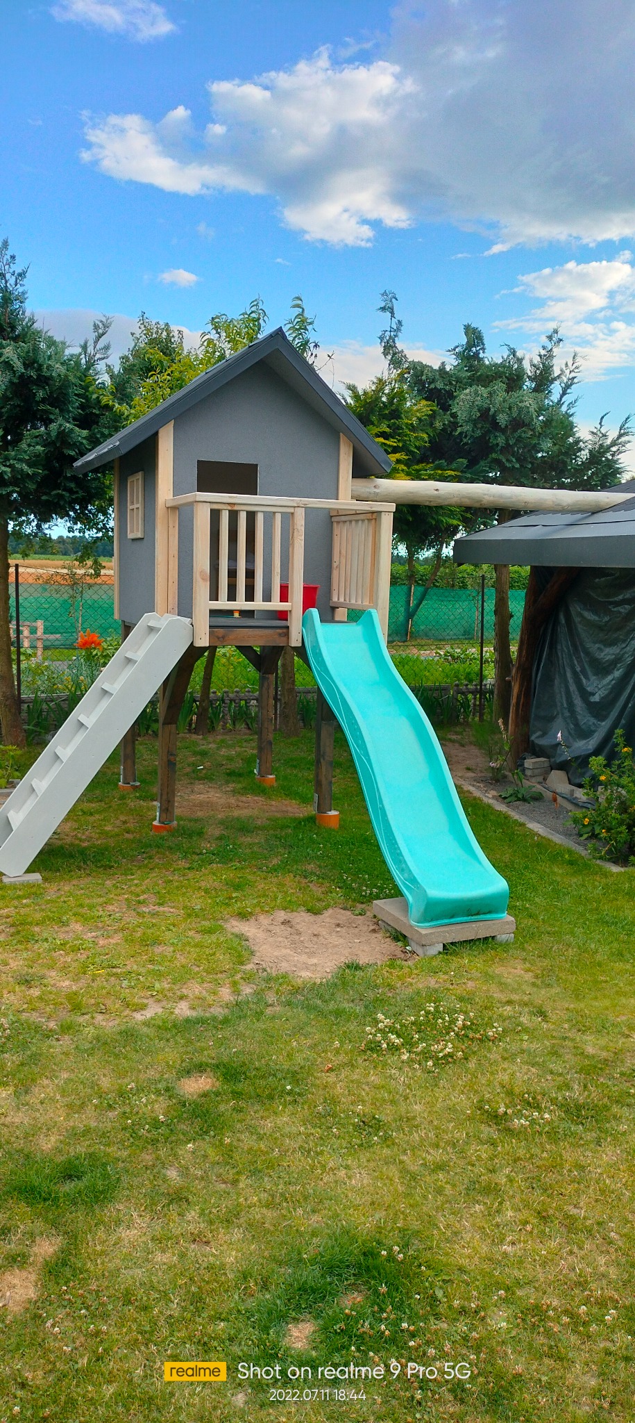 Drewniany domek dla dzieci z szarą elewacją, drabinką i turkusową zjeżdżalnią, umieszczony na trawniku w ogrodzie, z widoczną konstrukcją zadaszenia w tle.