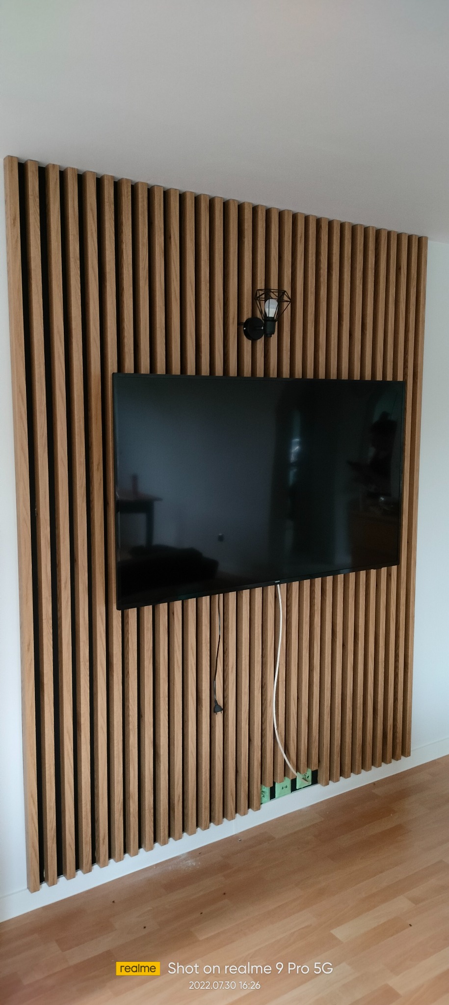 Drewniana ściana z pionowych listew, z zamontowanym telewizorem i lampą ścienną w stylu industrialnym, widoczne gniazdka elektryczne i przewody.