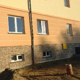 Dwukondygnacyjny budynek z elewacją w odcieniach beżu i brązu, z poziomymi paskami na górnej części i kamienną okładziną na dole, widok z poziomu gruntu, z widocznym wykopem w pobliżu fundamentów.