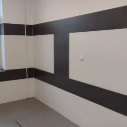 Wnętrze łazienki po remoncie, z ułożonymi białymi i grafitowymi płytkami w poziome pasy, widoczne okno z zamgloną szybą i szara podłoga.