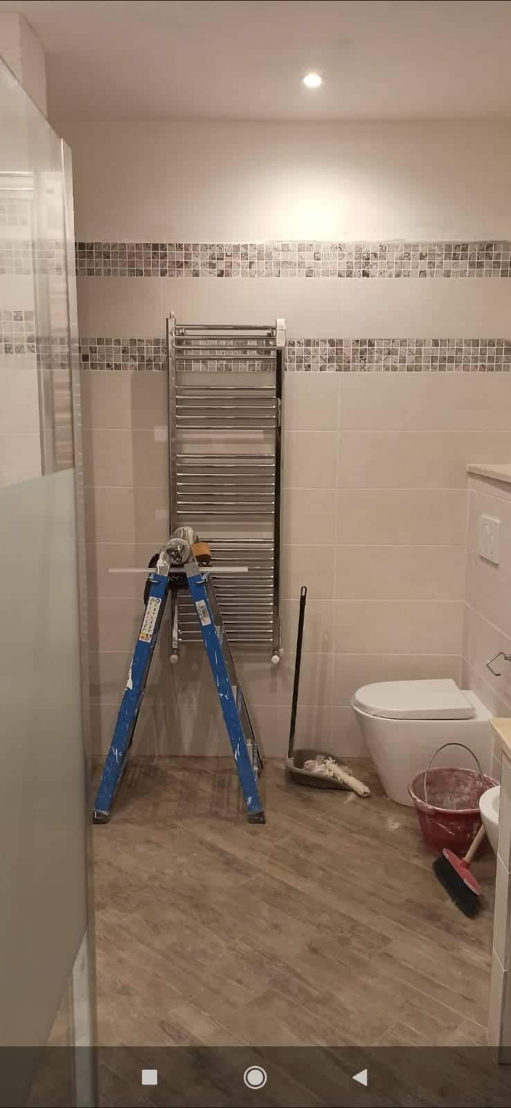 Łazienka w trakcie remontu: położone płytki imitujące drewno na podłodze, na ścianach jasne płytki z dekoracyjnym pasem mozaiki, drabinka grzejnikowa, niebieska drabina, wiadro z zaprawą i narzędzia.