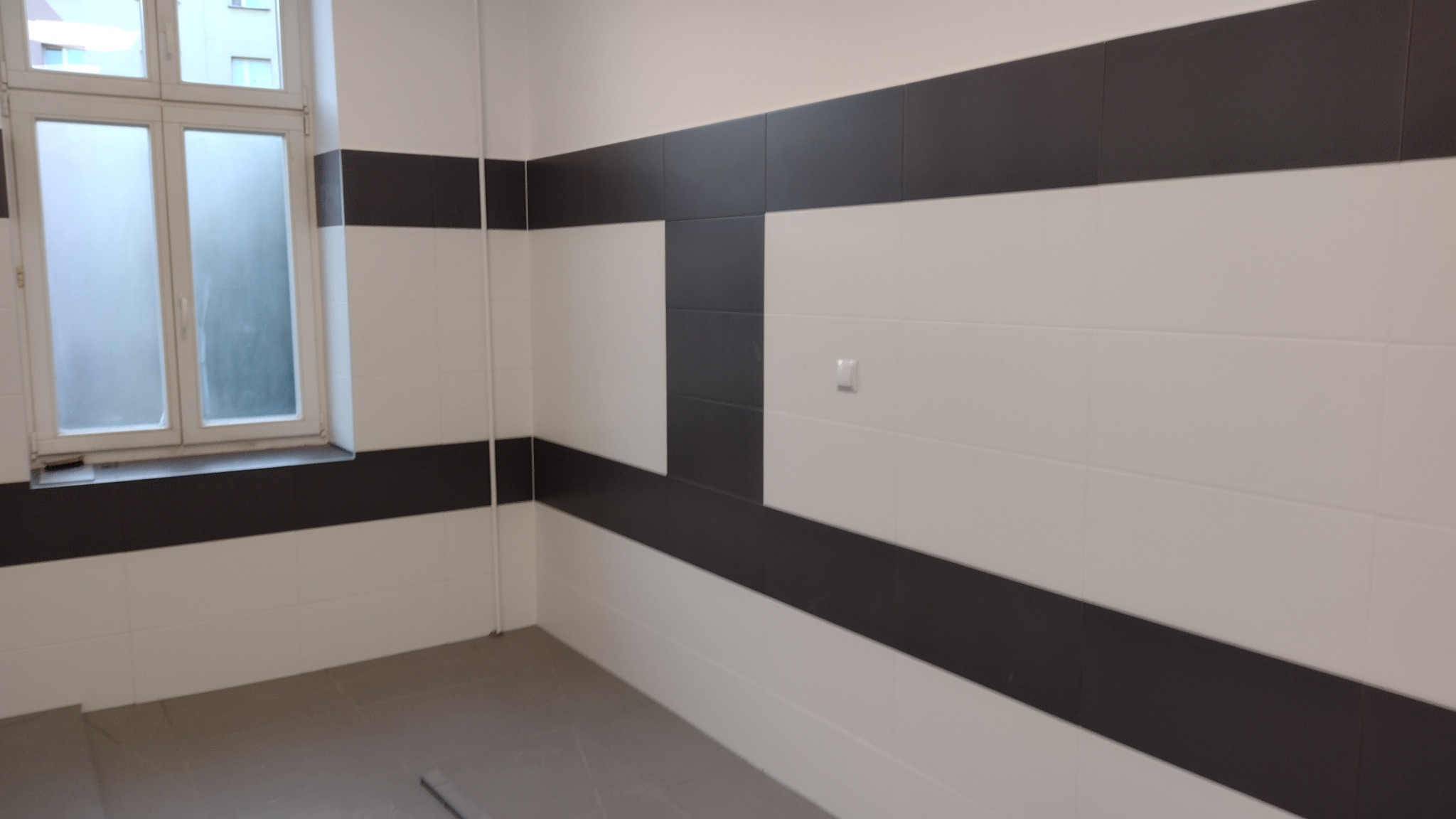 Wnętrze łazienki po remoncie, z ułożonymi białymi i grafitowymi płytkami w poziome pasy, widoczne okno z zamgloną szybą i szara podłoga.