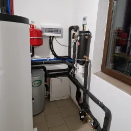 Zestaw urządzeń w kotłowni: biały zasobnik ciepłej wody, czerwony zbiornik wyrównawczy, czarny akumulator ciepła z izolowanymi rurami, szafka sterownicza na ścianie, instalacja na kafelkach.