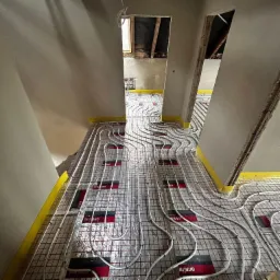 Białe rury ogrzewania podłogowego ułożone na folii z siatką, przytwierdzone taśmą. Widok z perspektywy podłogi w trakcie montażu instalacji w remontowanym wnętrzu.