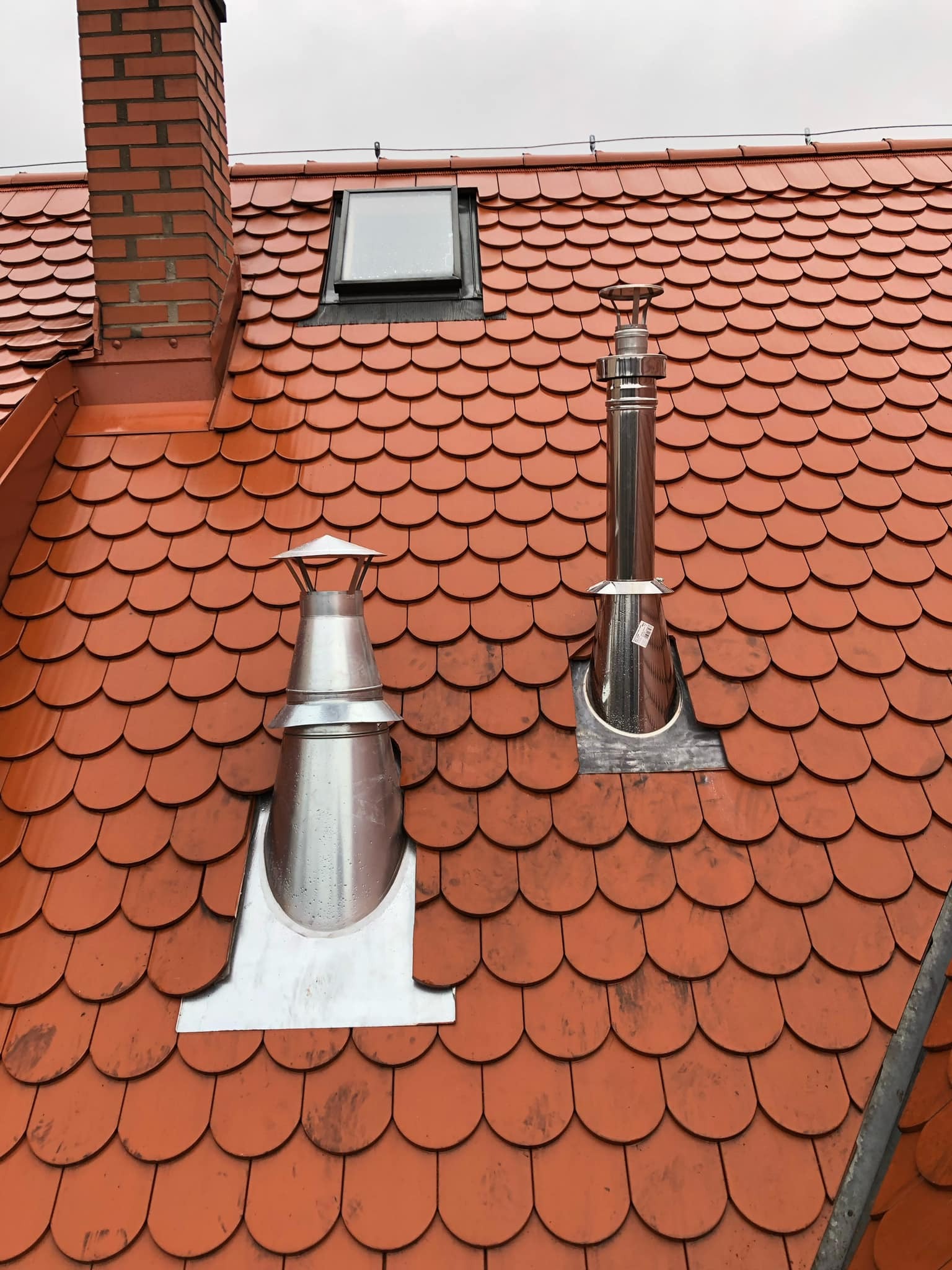 Czerwony dach z dachówki karpiówki z dwoma kominami ze stali nierdzewnej i oknem dachowym w pochmurny dzień.