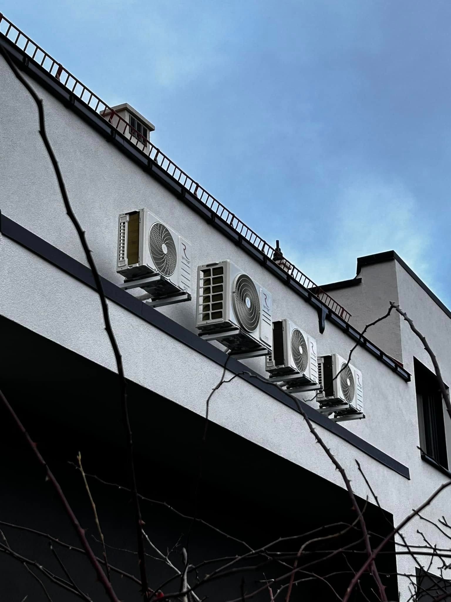 Biała fasada budynku z rzędem czterech zamontowanych klimatyzatorów na czarnej, poziomej listwie pod dachem, widok z dołu na tle pochmurnego nieba.