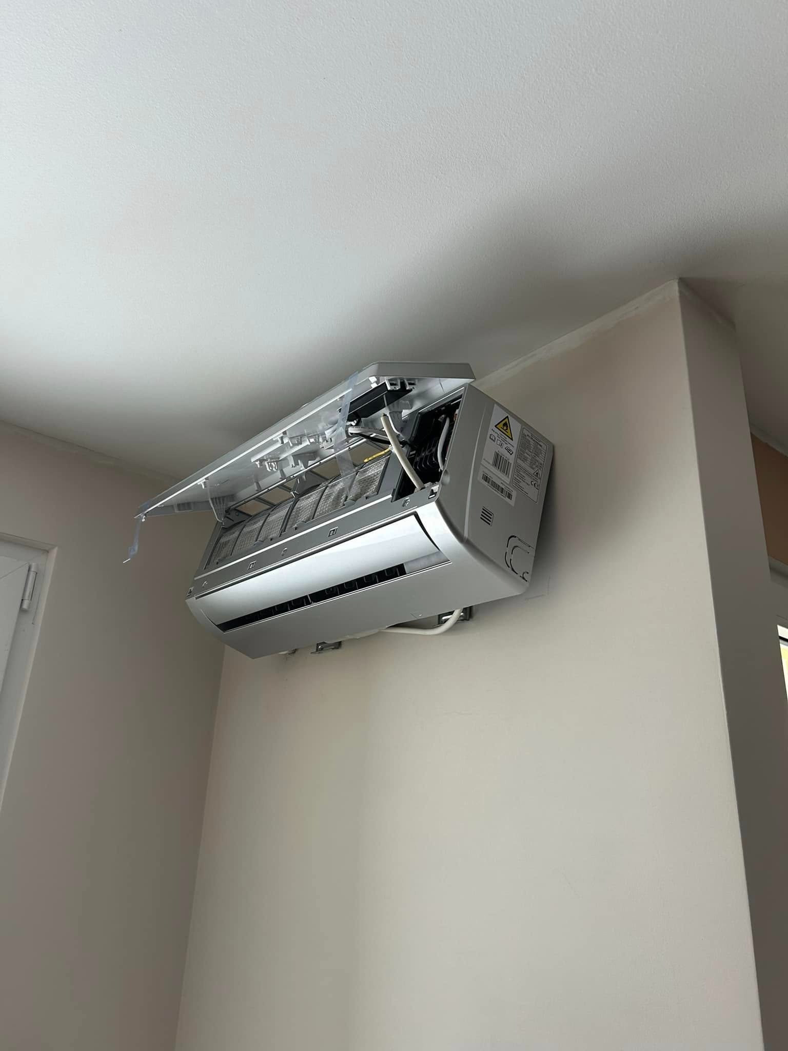 Ścienna klimatyzacja z otwartą obudową, ukazująca filtry i wewnętrzne elementy. Widoczne naklejki ostrzegawcze na boku urządzenia. Montaż w pomieszczeniu o jasnych ścianach.