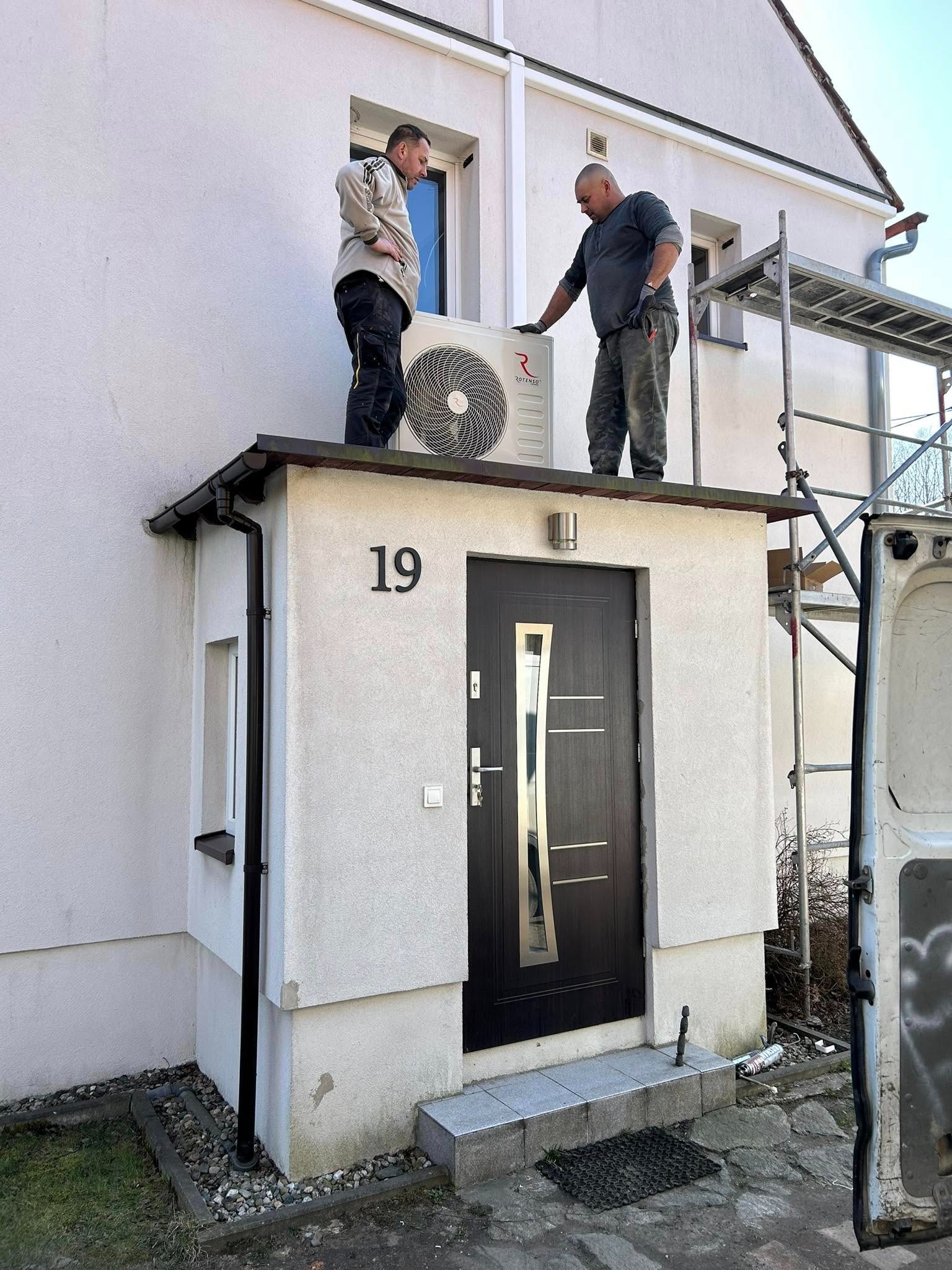 Dwóch monterów instaluje jednostkę zewnętrzną klimatyzacji Rotenso na daszku nad wejściem do domu z numerem 19. Widoczne rusztowanie i fragment samochodu dostawczego.