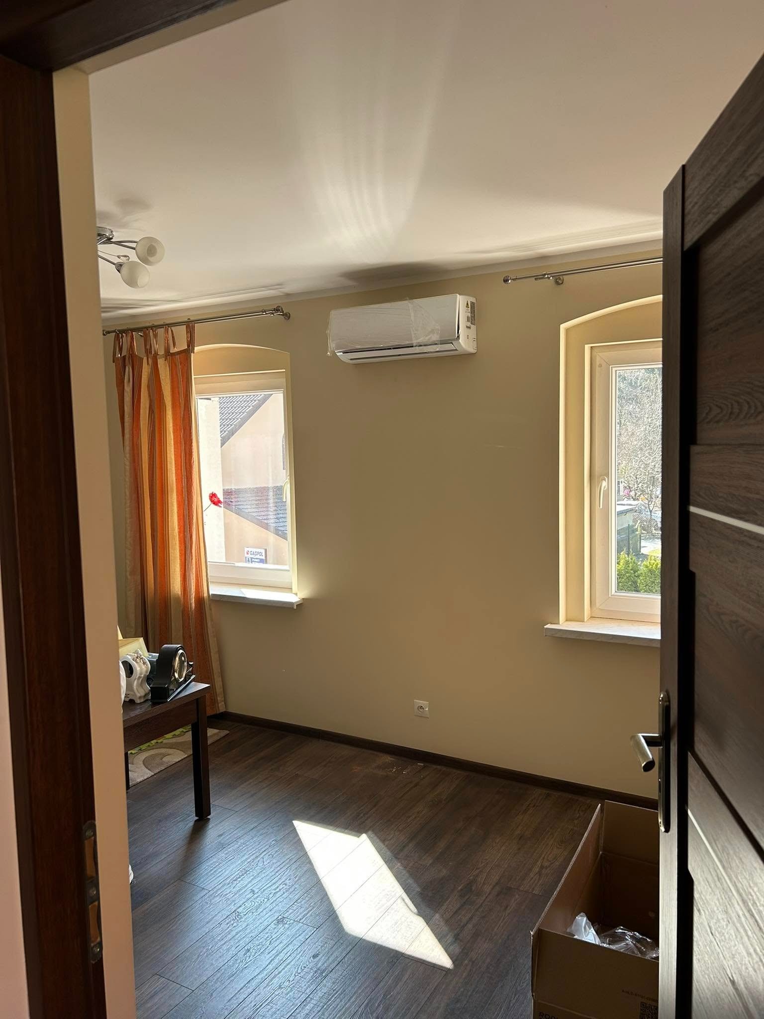 Wnętrze pokoju z zamontowaną klimatyzacją na ścianie, widok zza otwartych, ciemnych drzwi. Okna z jasnymi ramami, zasłonięte częściowo pomarańczowymi zasłonami.