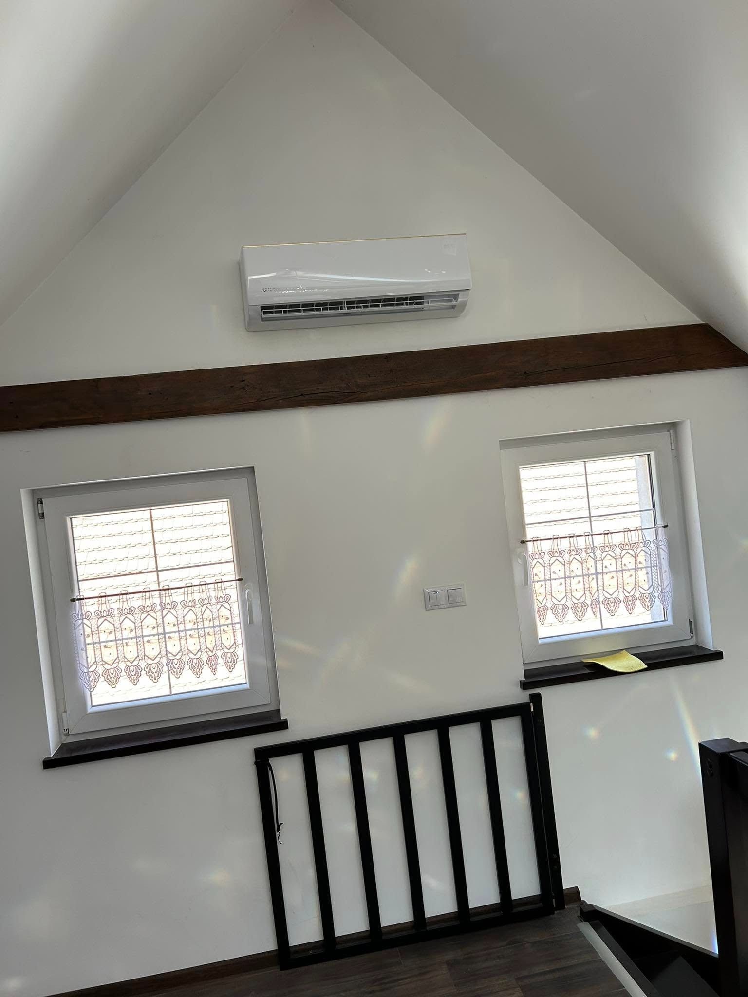 Klimatyzator na poddaszu z drewnianą belką, dwoma oknami z koronkowymi firankami i czarną barierką. Widoczne załamania światła.