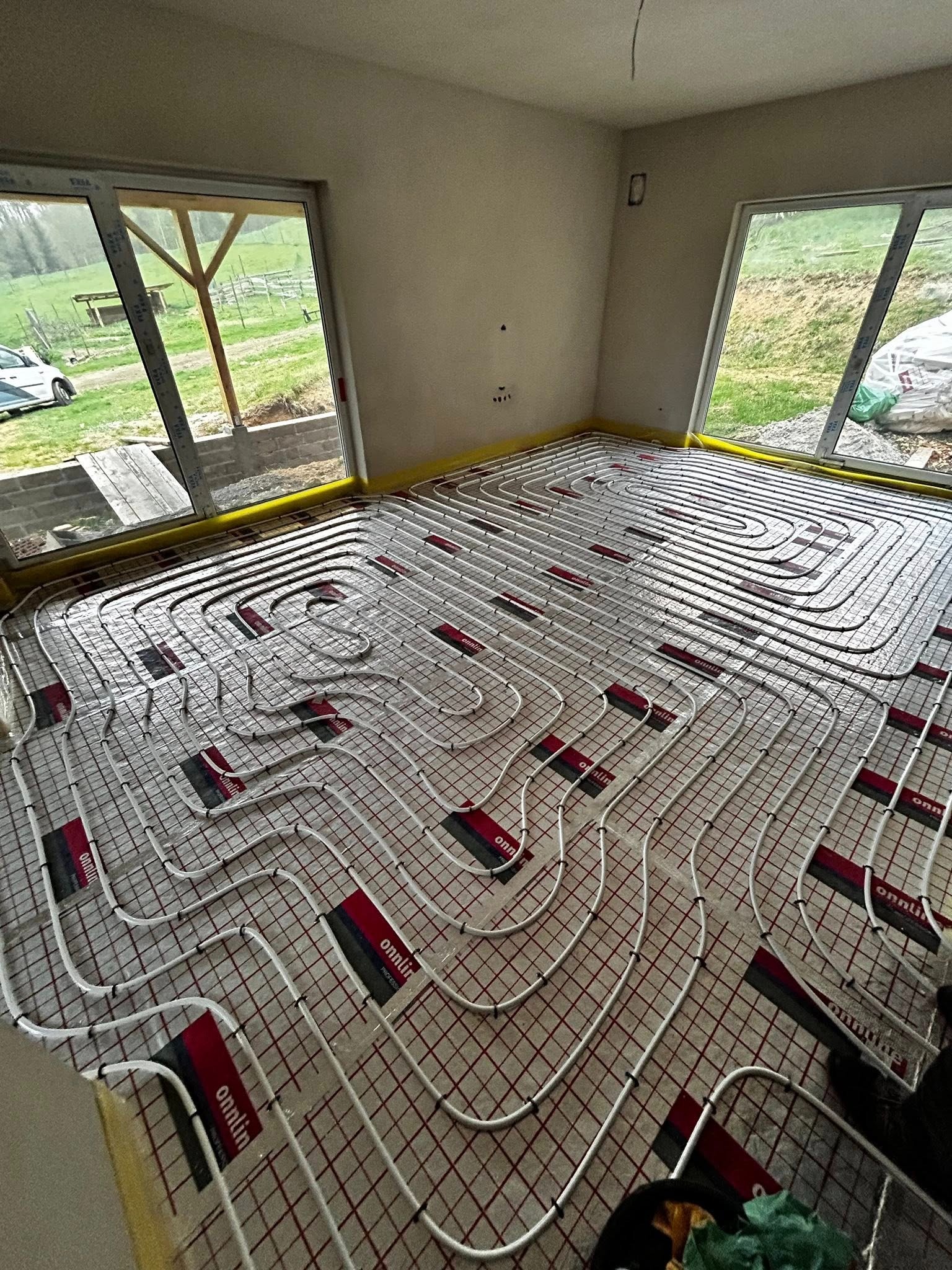 Rozłożona instalacja ogrzewania podłogowego z rurkami na siatce, widok z góry w surowym wnętrzu z oknami na zewnątrz. Żółta taśma na ścianach.