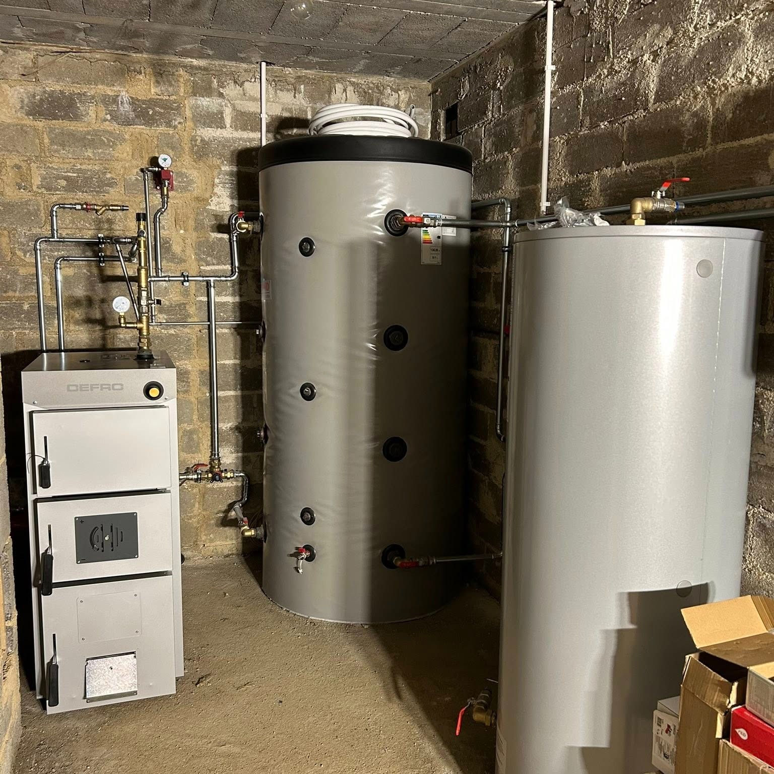 Szary piec Defro z metalowymi rurami i dwa szare zbiorniki na wodę w pomieszczeniu z ceglanymi ścianami. Elementy instalacji hydraulicznej.