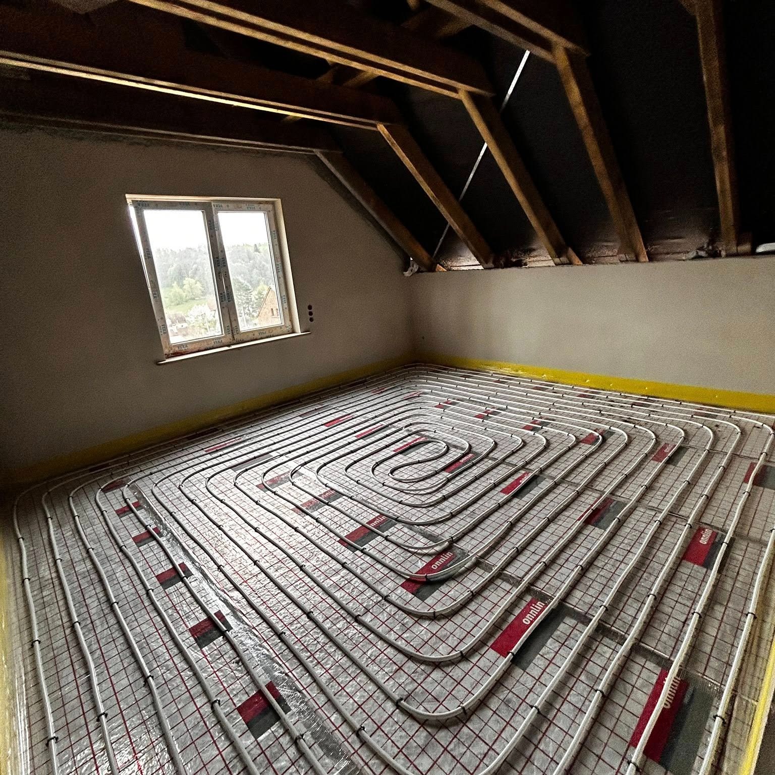 Instalacja ogrzewania podłogowego w poddaszu z oknem. Białe rurki ułożone w spiralny wzór na srebrnej macie, widoczne elementy drewnianej konstrukcji dachu.