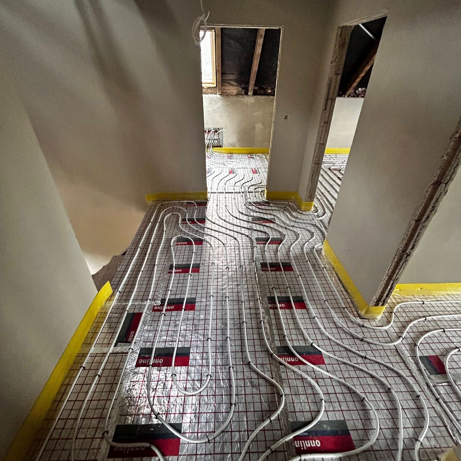 Białe rury ogrzewania podłogowego ułożone na folii z siatką, przytwierdzone taśmą. Widok z perspektywy podłogi w trakcie montażu instalacji w remontowanym wnętrzu.