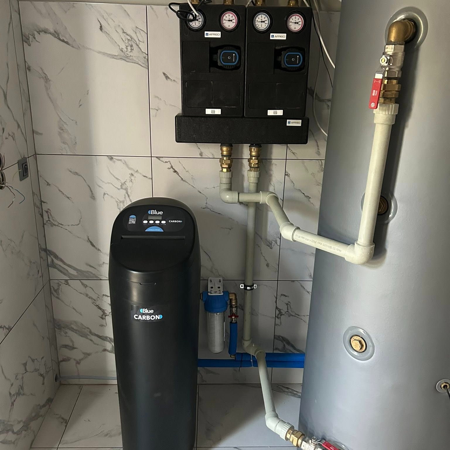 Instalacja uzdatniania wody z zbiornikiem i dwoma czarnymi modułami Afriso na marmurowej ścianie. Widoczne białe i niebieskie rury oraz zawory.