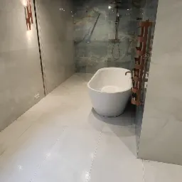 Nowoczesna łazienka z wolnostojącą białą wanną, marmurowymi płytkami na ścianach i podłodze oraz dekoracyjnym grzejnikiem drabinkowym w kolorze miedzi.