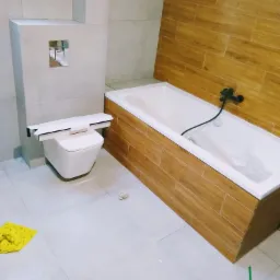 Nowoczesna łazienka w trakcie remontu: podwieszana toaleta z zabudową, wanna obudowana płytkami imitującymi drewno, widoczne niedokończone prace na suficie, żółta ścierka i wiadro na podłodze.