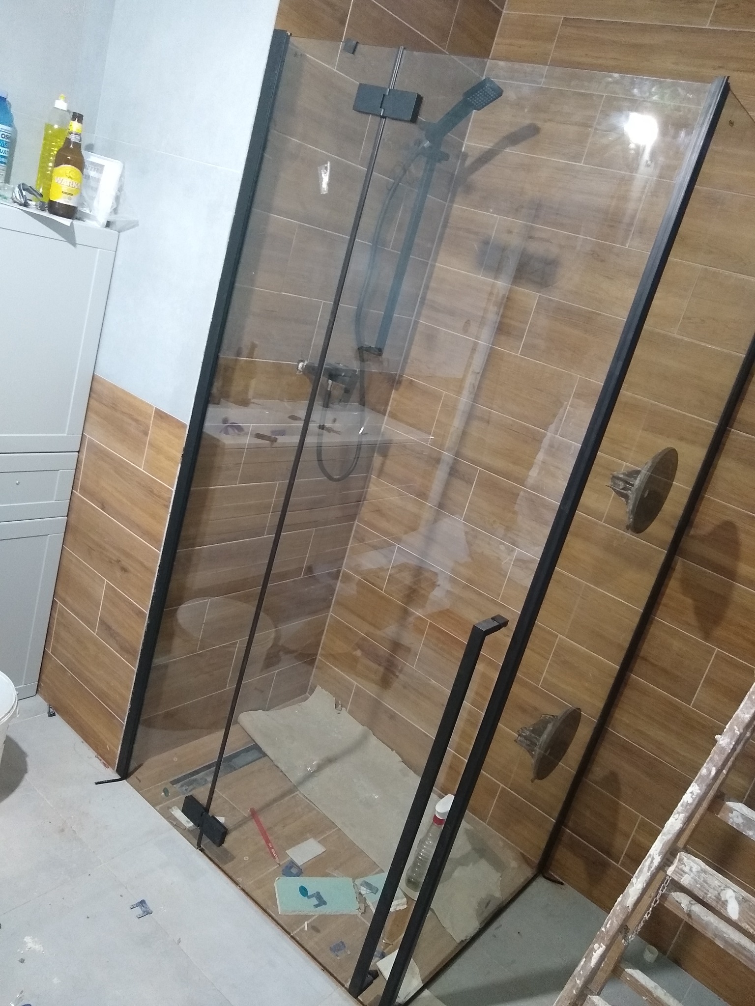 Montaż kabiny prysznicowej typu walk-in z czarnymi profilami, widoczne płytki imitujące drewno na ścianach, niedokończone prace wykończeniowe na podłodze i drabina malarska w tle.