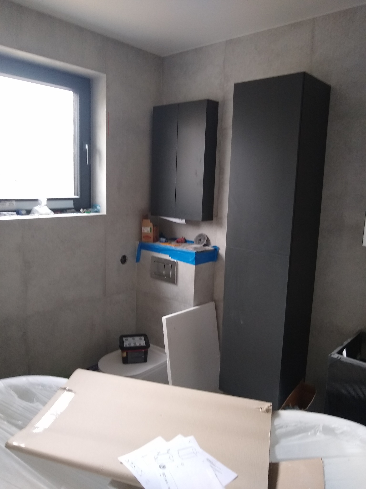 Wnętrze łazienki w trakcie prac remontowych, widoczne szare płytki, czarne szafki wiszące, zabudowana wanna w folii ochronnej oraz kartony z dokumentacją na wierzchu.