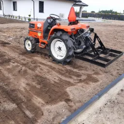 Pomarańczowy traktor Kubota z zamontowanym narzędziem do wyrównywania gleby na rozległym, przygotowanym terenie pod trawnik, widoczny obrys krawężnika, w tle biały dom z ciemnymi oknami.