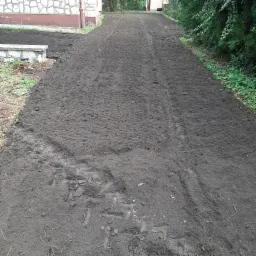 Przygotowany teren pod trawnik z rolki, świeżo wyrównana ziemia z widocznymi śladami opon, budynek w tle.