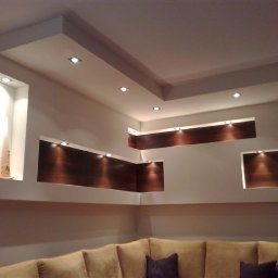 Robert Góra - Nowoczesna aranżacja ściany z wnękami podświetlanymi punktowym oświetleniem, z elementami dekoracyjnymi z ciemnego drewna i gładzią gipsową, widoczny fragment sofy.