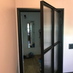 Robert Góra - Otwarta, dwuskrzydłowa, czarna, przeszklona drzwi wewnętrzne z ryflowanym szkłem, widoczne z boku, z widokiem na odnowione wnętrze z drewnianą podłogą i lustrem.