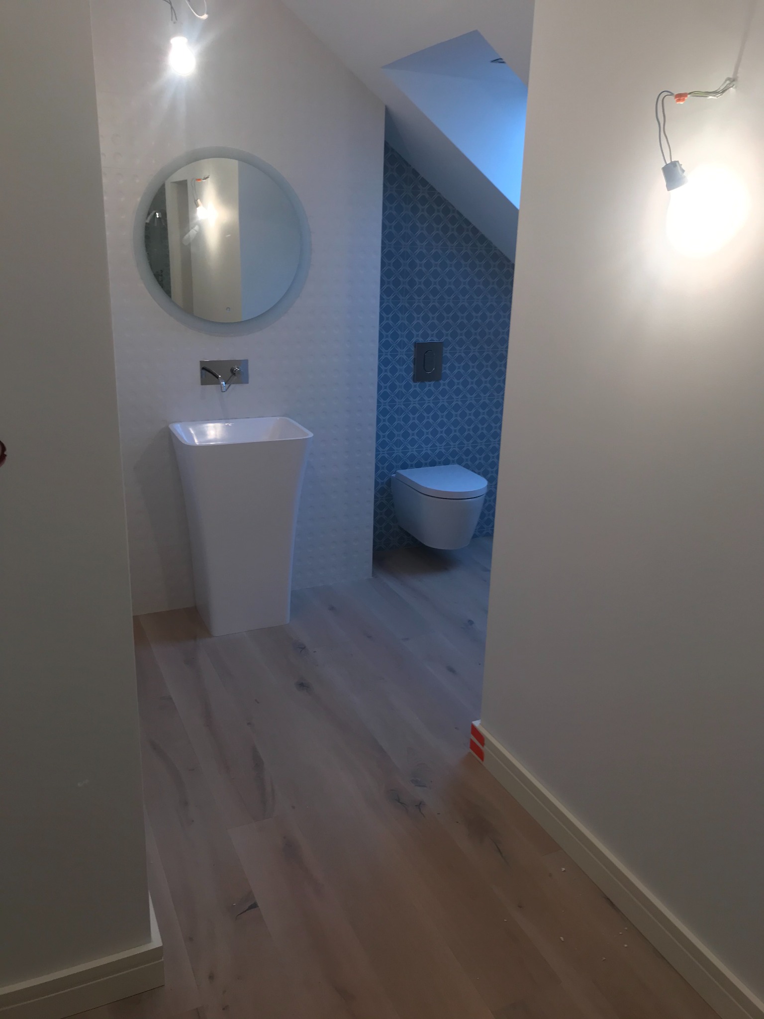 Nowoczesna łazienka w trakcie wykańczania: wolnostojąca umywalka, okrągłe lustro, toaleta w tle, drewnopodobna podłoga, niebieska mozaika na ścianie wnęki, surowa żarówka.