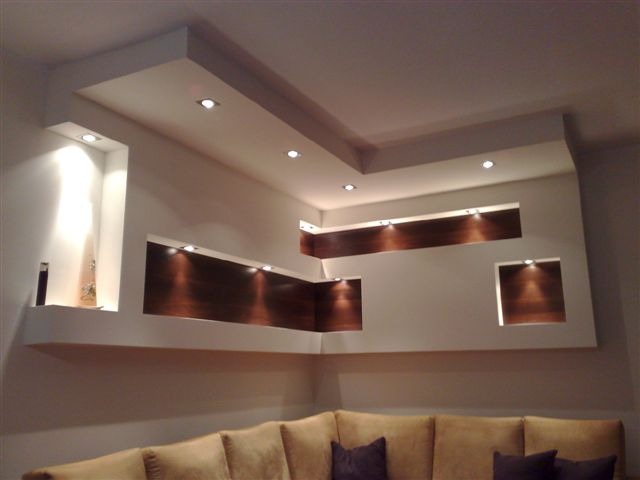 Nowoczesna aranżacja ściany z wnękami podświetlanymi punktowym oświetleniem, z elementami dekoracyjnymi z ciemnego drewna i gładzią gipsową, widoczny fragment sofy.