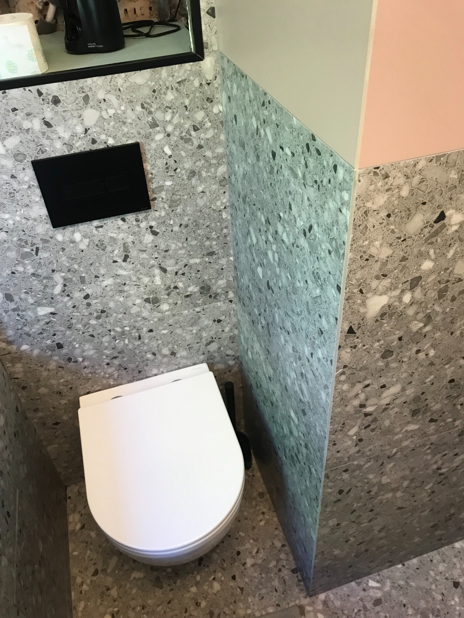 Nowoczesna toaleta z misą wiszącą, obudowana płytkami lastryko w odcieniach szarości, z czarnym panelem spłukującym i fragmentem ściany pomalowanej na różowo i beżowo.