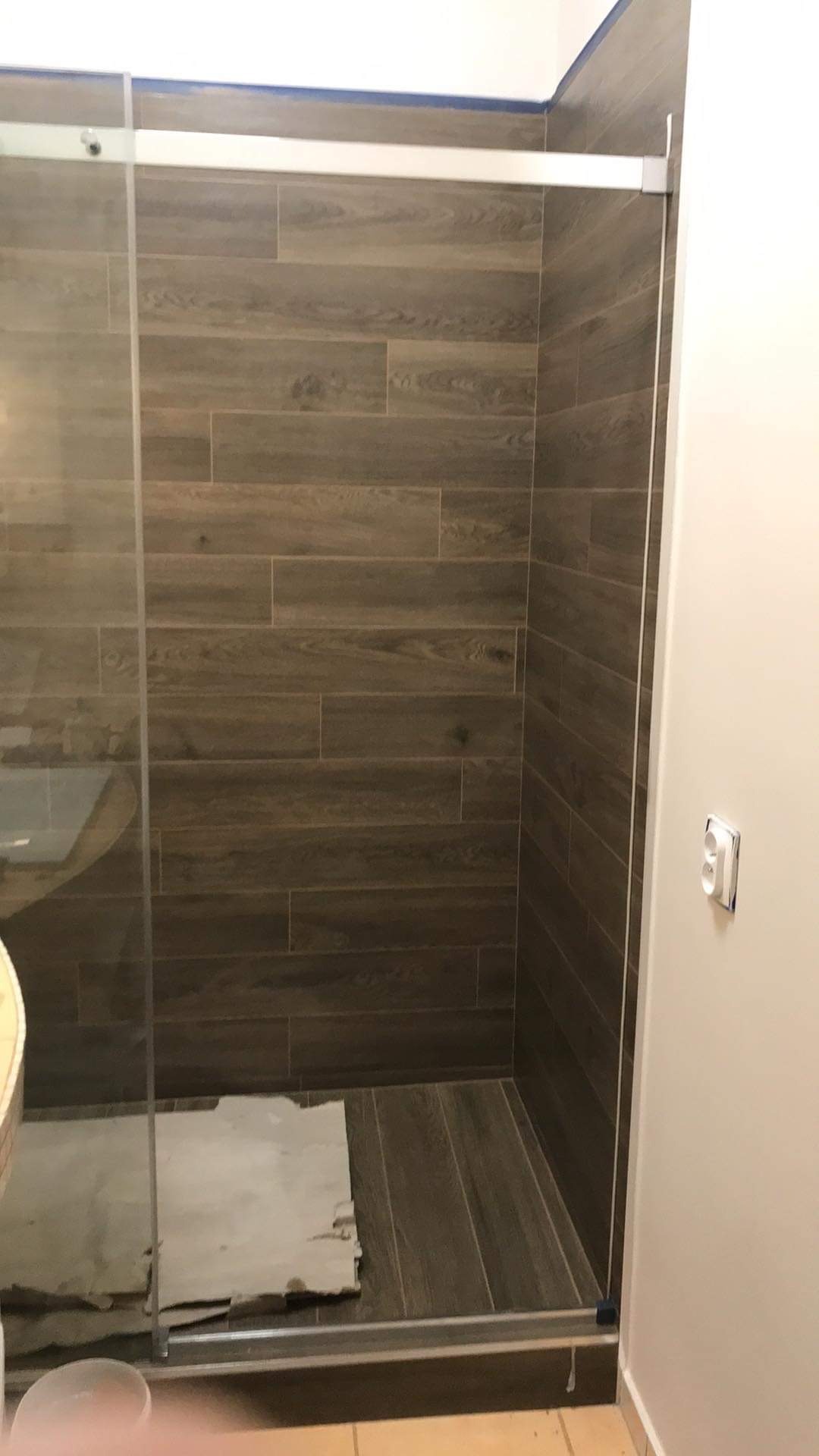 Nowoczesna kabina prysznicowa z przesuwnymi drzwiami, wykończona brązowymi płytkami imitującymi drewno na ścianach i podłodze. Widoczny fragment umywalki i zabezpieczenie podłogi wewnątrz kabiny.