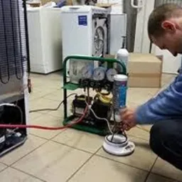 Mężczyzna w szarym swetrze podłącza przewody do sprężarki lodówki za pomocą specjalistycznego urządzenia z manometrami. W tle inne sprzęty AGD.