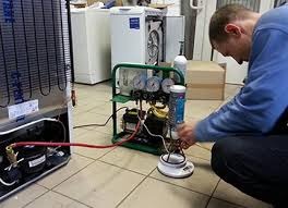 Mężczyzna w szarym swetrze podłącza przewody do sprężarki lodówki za pomocą specjalistycznego urządzenia z manometrami. W tle inne sprzęty AGD.