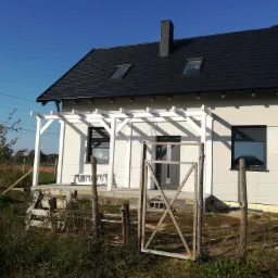 Dom jednorodzinny z czarnym dachem z dachówki, białymi oknami i białą pergolą, widoczny zza prowizorycznego ogrodzenia z siatki.